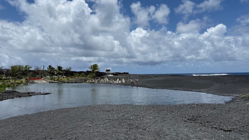 Looking back from Pohoiki black sand beach toward the lifeguard tower, warm thermal pond, parking area, and 2018 lava flow at Isaac Hale Beach Park