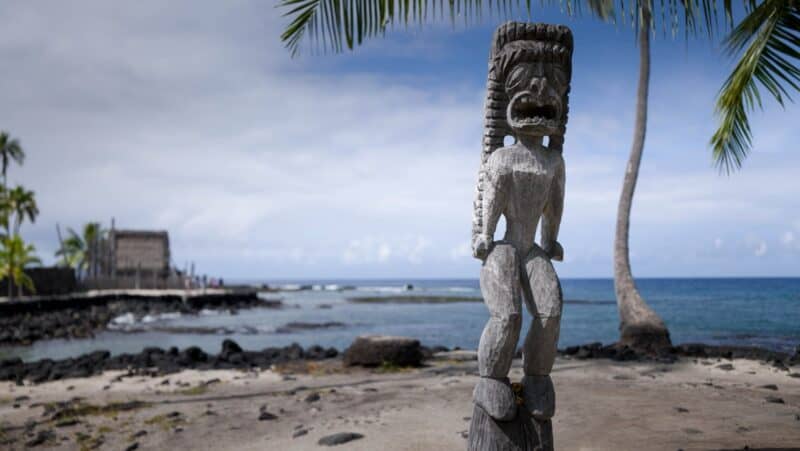 A carved wooden kiʻi image at Puʻuhonua o Hōnaunau, with the Hale o Keawe temple and the bay behind