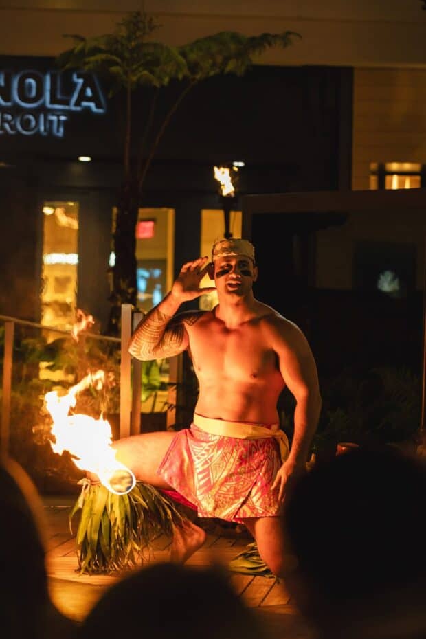 Fire knife dancer beside a lit torch with a shopfront and a red exit sign behind him at Queens Waikiki Luau