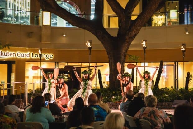 Hula dancers raising wooden canoe paddles beneath the courtyard tree at Queens Waikiki Luau