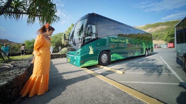 Green Roberts Hawaii motorcoach parked at Hanauma Bay with a staff member in an orange muʻumuʻu waiting beside it