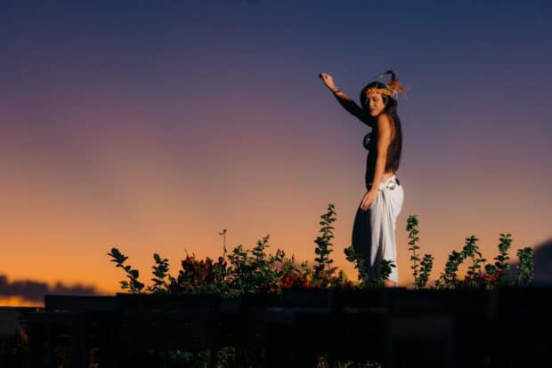 Silhouetted hula dancer in a white pāʻū skirt performing against a dusk sky at the Myths of Maui Luau