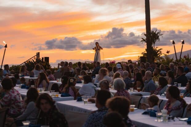 Guests in leis at long dinner tables watching a hula dancer as the sun sets over Kāʻanapali