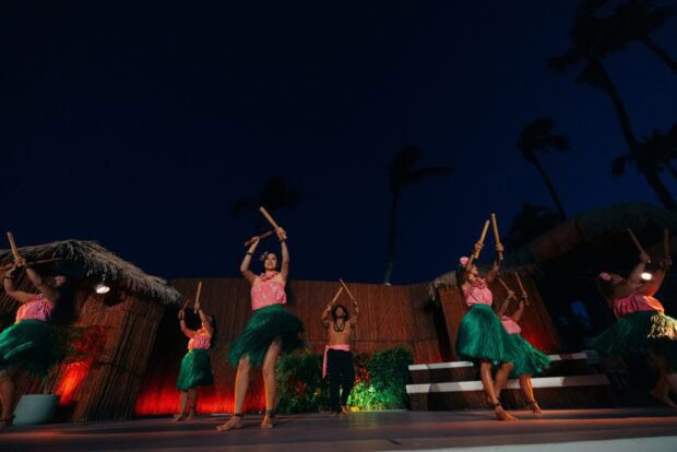Group of dancers in ti-leaf skirts performing with bamboo puʻili sticks on the lit stage at the luau