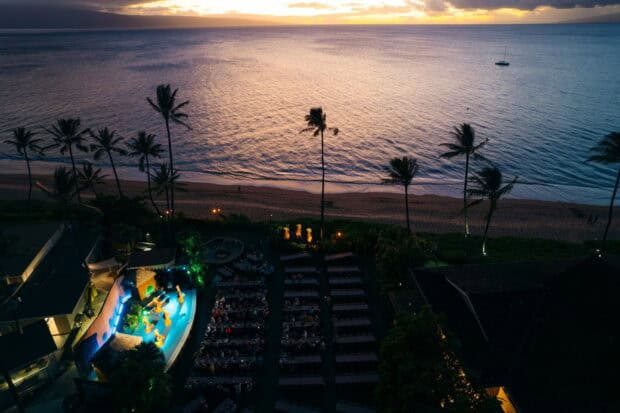 Aerial view of the Myths of Maui Luau grounds at dusk with lit stage, dinner tables, palms, and Kāʻanapali Beach behind