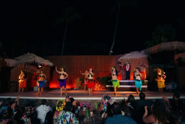 Line of dancers in coconut-shell tops and colored skirts performing Tahitian dance as the audience watches