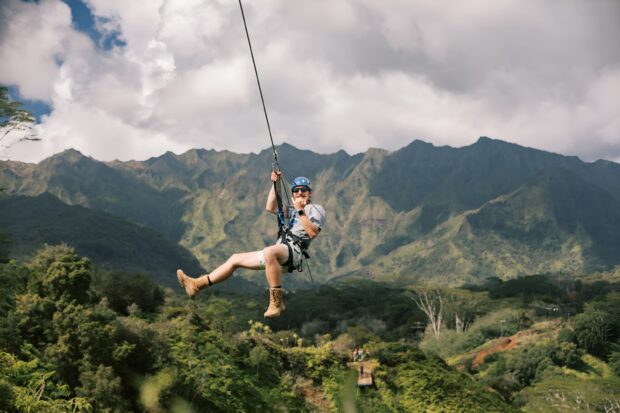 Rider throwing a shaka while ziplining toward a landing platform with Kauai mountains in the background