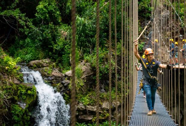 Guest in an orange helmet crossing a steel suspension bridge next to a waterfall in the rainforest
