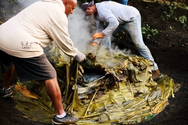 Two hosts pull back steaming banana leaves to uncover foil trays of food in the imu at Imu Mea ʻAi