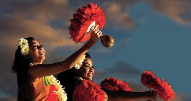 Waikīkī Starlight Luau dancers holding red-feathered gourd rattles, lit by low sun against a cloudy sky