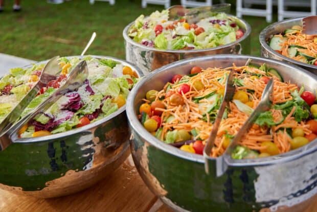 Four stainless bowls of green salad with tomatoes, cucumber and shredded carrot, tongs resting in each