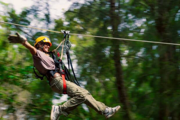 Zipline rider in a yellow helmet with one arm outstretched, forest blurring past at speed