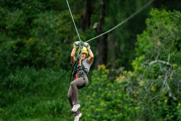 Rider in a yellow helmet seated in the harness mid-zipline against green rainforest in North Kohala