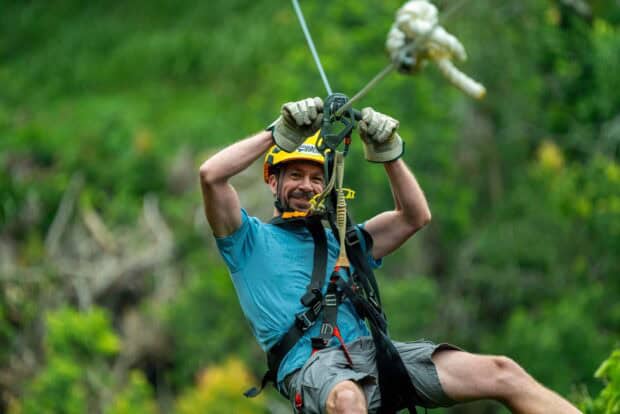 Smiling man in a teal shirt and yellow helmet gripping the trolley while ziplining through green forest